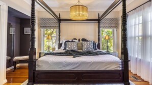 A four-poster canopy bed with a chandelier, a tufted headboard, and a view of greenery outside.