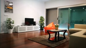 A modern living room with a flat-screen TV on a white cabinet, a coffee table with a plant, a beige sofa, and a large window with blinds.