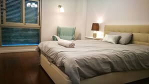 A neatly made bed with a rolled towel, a green chair, a bedside lamp, and a window with blinds.