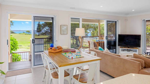 A spacious living room with a wooden table, white chairs, a beige sofa, and a television. Large windows offer a view of a grassy area and trees. - Allure by the Sea - beachfront luxury home (Bonny Hills)