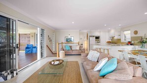 A modern living room with a glass coffee table, a beige sofa with patterned cushions, and a dining area with a wooden table and white chairs. - Allure by the Sea - beachfront luxury home (Bonny Hills)