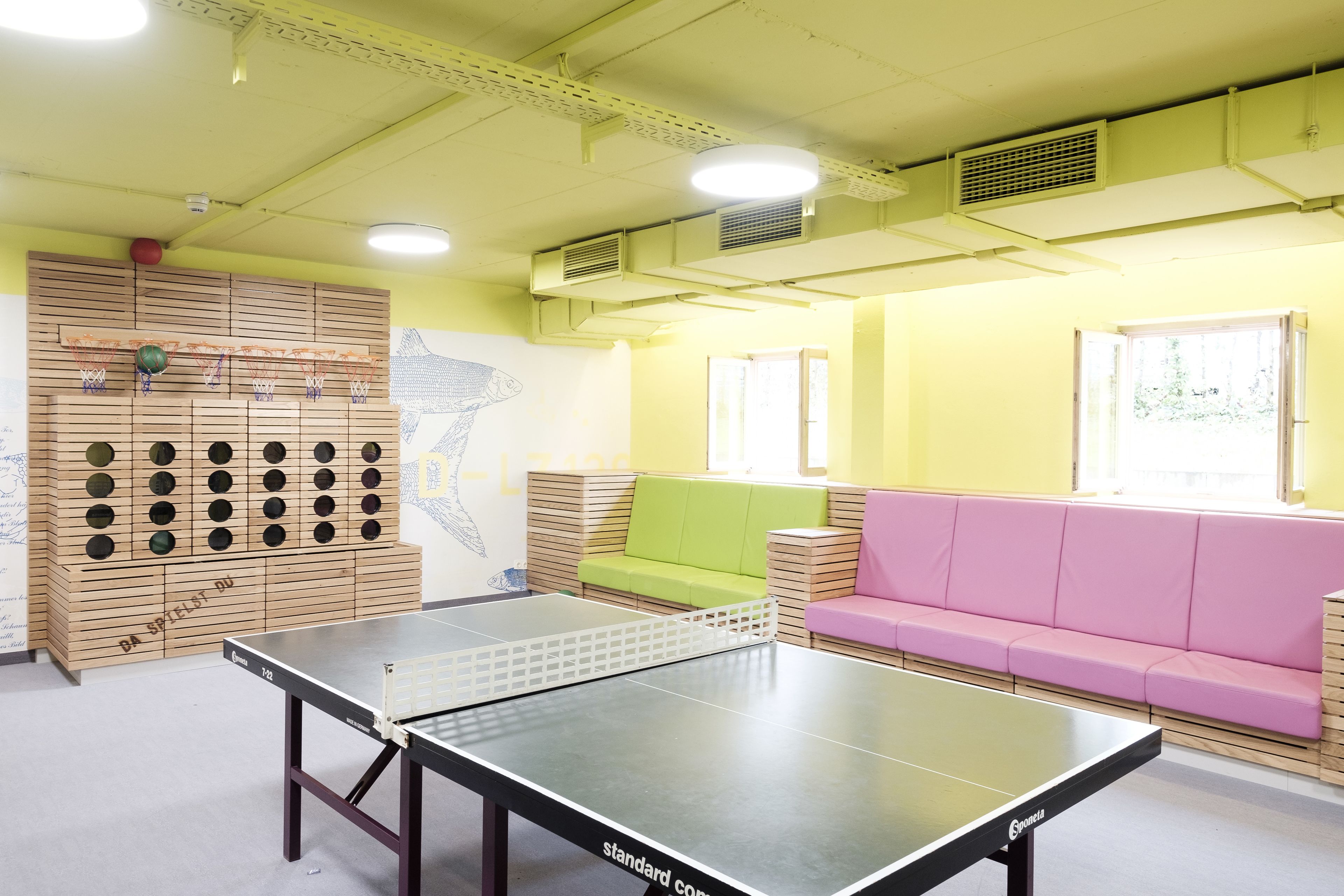 Una mesa de ping pong en una habitación con pared amarilla, un estante de madera con pelotas y un sofá rosa.
