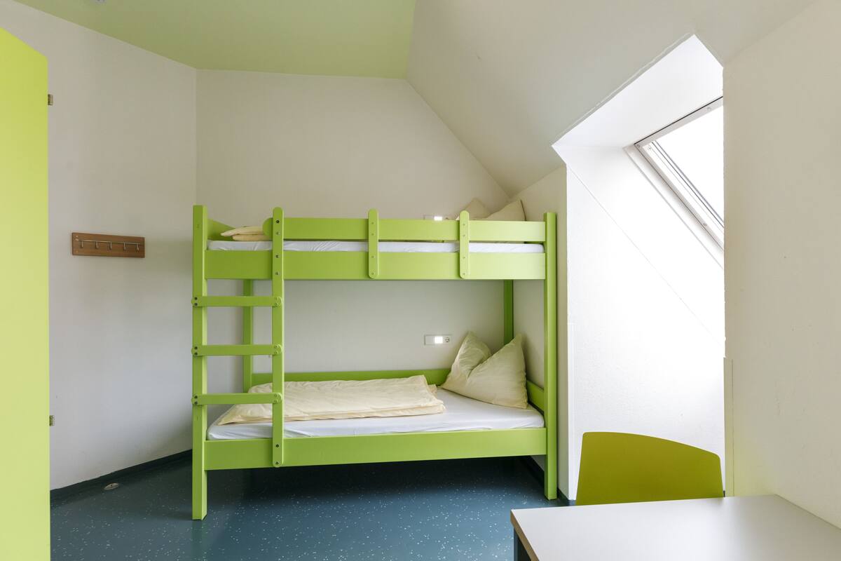 Ein Stockbett mit einer Leiter, ein kleiner Tisch und ein Fenster in einem Zimmer mit geneigter Decke.