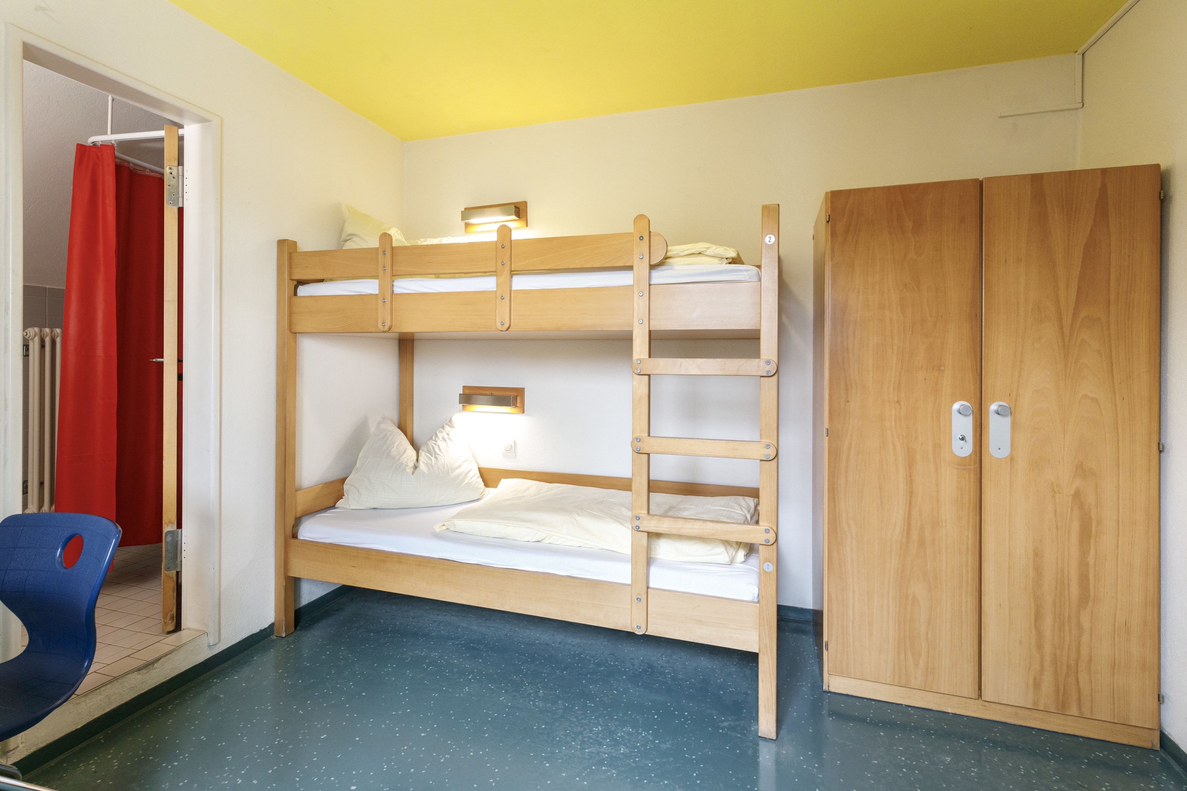Een slaapkamer met een stapelbed, een houten garderobe, een stoel en een rood gordijn.