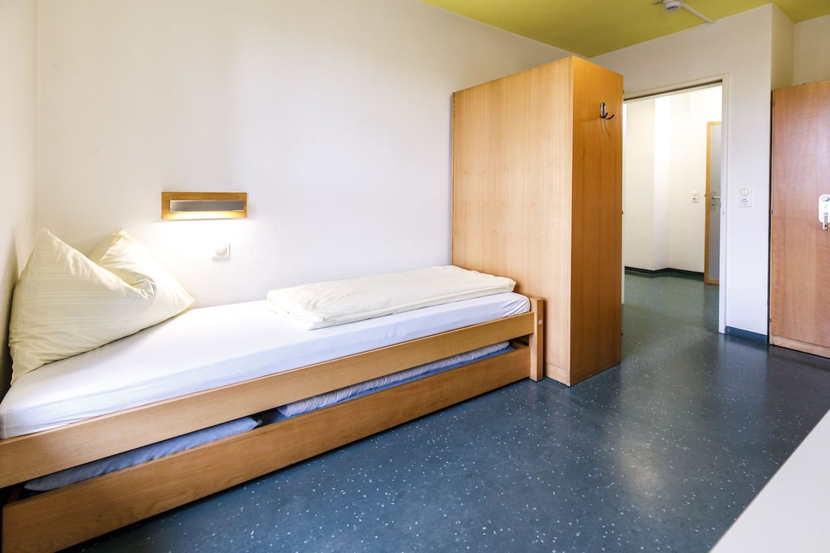 Ein Zimmer mit einem Einzelbett, einem Holzbettrahmen, einem kleinen Nachttisch und einer Wandleuchte.
