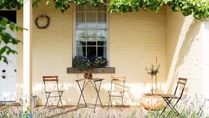 A small outdoor seating area with two wooden chairs and a table, a window with a flower box, and a wall-mounted wreath. - Cosy Heritage Cottage in Newstead, Central to Daylesford, Maldon & Castlemaine (Newstead)