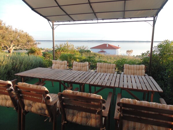 A covered outdoor seating area with a table and chairs, overlooking a body of water and a house in the background.