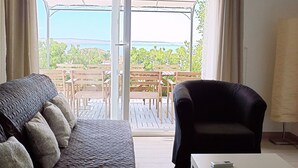 Een kamer met een bank, een stoel, een bijzettafeltje en uitzicht op een terras met tafels en stoelen.