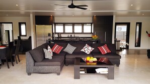 A modern living room with a dark grey sofa, a coffee table with fruits, and a kitchen in the background. - Boaza Place is situated in Nikao and offers an outdoor swimming pool.  (Puapuautu Tapere)
