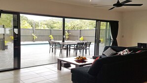 A modern living room with a sofa, a coffee table, and a dining area with chairs and a table. There is a sliding glass door leading to an outdoor patio with a pool and a view of trees and a fence. - Boaza Place is situated in Nikao and offers an outdoor swimming pool.  (Puapuautu Tapere)