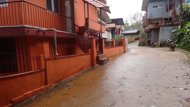 A residential street with houses, a visible water drain, and a motorcycle parked on the side. - For delightful stay, visit us (Madikeri)