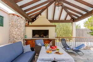 Une terrasse extérieure avec un mur en pierre, un barbecue, un canapé, une table à manger avec de la nourriture et deux chaises longues.