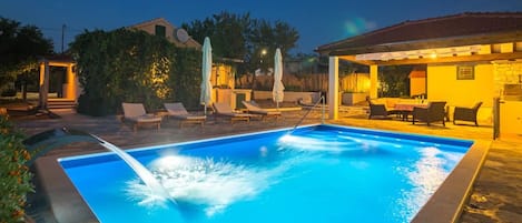 A swimming pool with a water feature, surrounded by lounge chairs and umbrellas, with a building in the background.