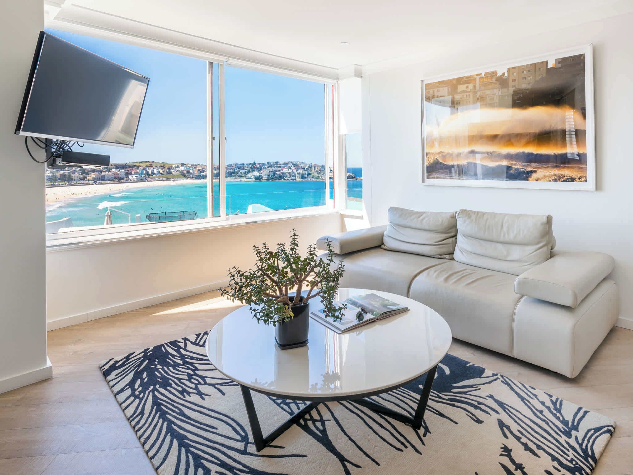 A modern living room with a sofa, a coffee table with a plant, a TV mounted on the wall, and a large window with a city view.