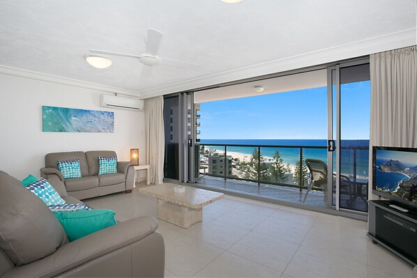 Relax in this stylish living room with a sofa, flat-screen TV, and coffee table, opening up to a balcony with ocean and city views.