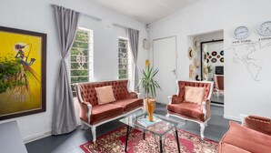 A living room with a red sofa, a glass coffee table, and a red patterned rug. There are two windows with curtains, a painting on the wall, and a map of the world. - Modern Bohemian Apartment with Pool, Fibre Wi-Fi and Bbq Patio in Centurion (Centurion)