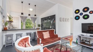 A modern living room with a sofa, a coffee table, and a television. There is a kitchen area with a bar counter and stools. The room has a large window with curtains and a wall with a chalkboard. - Modern Bohemian Apartment with Pool, Fibre Wi-Fi and Bbq Patio in Centurion (Centurion)