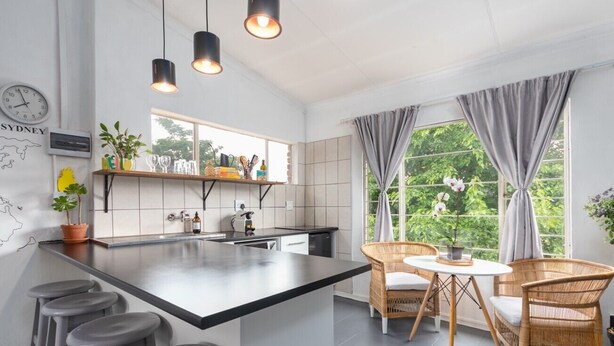 A modern kitchen with a central island, bar stools, wicker chairs, and a view of greenery through large windows. - Modern Bohemian Apartment with Pool, Fibre Wi-Fi and Bbq Patio in Centurion (Centurion)