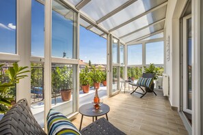 Uma varanda espaçosa com grandes janelas, uma mesa com garrafa e copos, e uma cadeira.