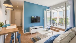 A modern living room with a blue accent wall, a flat-screen TV, a dining area with a wooden table and blue bar stools, and a balcony with outdoor furniture. - Enjoy your private terrace near City Park! (Budapest)