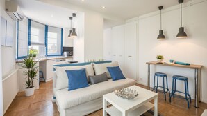 A modern living space with a white sofa, blue cushions, a small table, and a kitchen area with a countertop and stools. - Enjoy your private terrace near City Park! (Budapest)