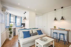 Un espace de vie moderne avec un canapé blanc, des coussins bleus, une petite table et un coin cuisine doté d’un comptoir et de tabourets.
