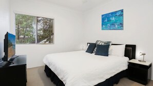 Ein Schlafzimmer mit Bett, Fernseher, Nachttisch, Lampe, einem Fenster mit Jalousien und einem Bild an der Wand.