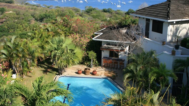 A pool surrounded by lush greenery, a white building with a tiled roof, and a view of boats on a bay.