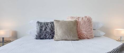 A neatly made bed with three pillows in different textures and colors, two bedside lamps, and a white wall.