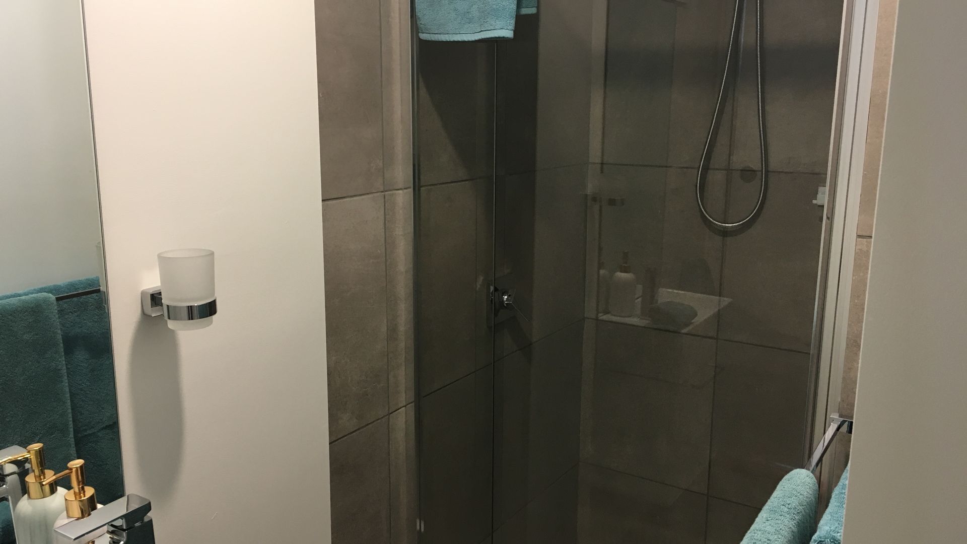 Et moderne badeværelse med et glasafskærmet brusebad, en hvid vægmonteret sæbedispenser og en lille vask med toiletartikler.