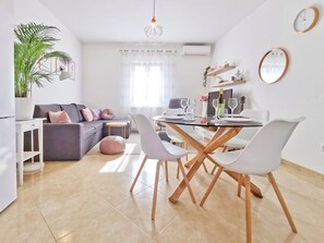 Un salon moderne avec un canapé gris, une table à manger entourée de chaises blanches, une plante en pot et un luminaire suspendu.