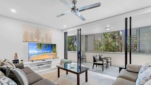 Un salon moderne doté d’un téléviseur à écran plat, d’un ventilateur de plafond et d’une grande fenêtre offrant une vue sur les arbres.