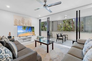 A modern living room with a flat-screen TV, a ceiling fan, and a large window with a view of trees.