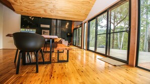 A modern interior with a wooden floor, a dining area with a table and chairs, and large windows offering a view of the outdoors.
