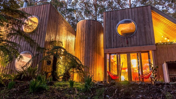 Una cabaña moderna de madera con ventanas circulares y una puerta de cristal que da a un interior con una silla roja.