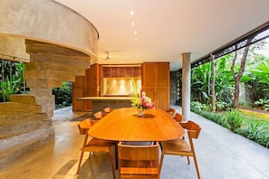 Un espace repas moderne avec une grande table et des chaises en bois, un îlot central doté d’un plan de travail en marbre et une vue sur une végétation luxuriante à l’extérieur.