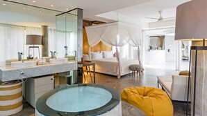 Una moderna camera d'albergo con un grande letto, un bagno con pareti in vetro, un'area salotto con un poggiapiedi giallo e uno specchio grande.