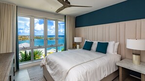 Habitación de hotel con una cama grande, un ventilador de techo, una mesita de noche con lámpara y vistas al mar a través de amplios ventanales.