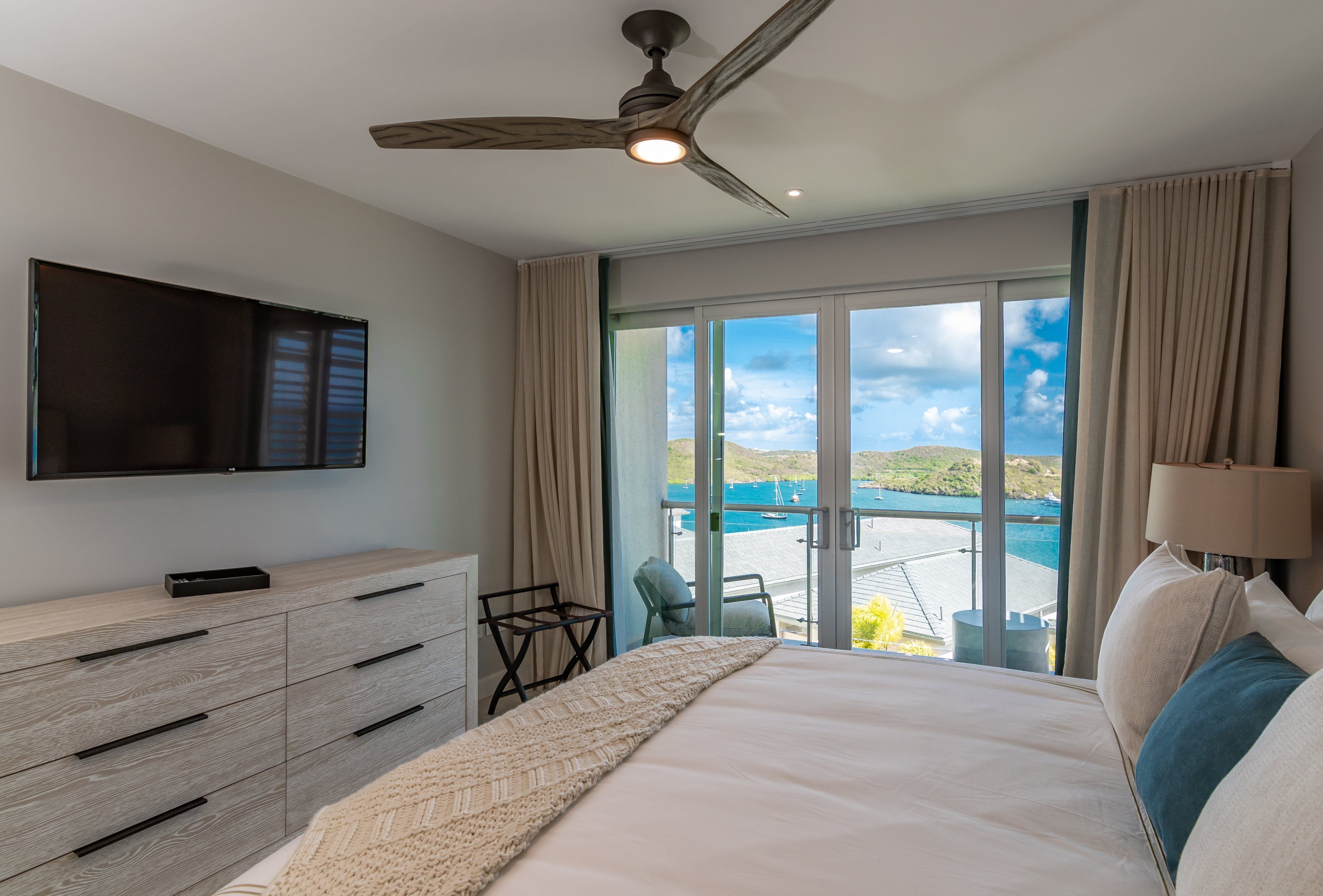 A hotel room with a bed, a TV, a ceiling fan, a view of the sea, and a balcony with a chair.