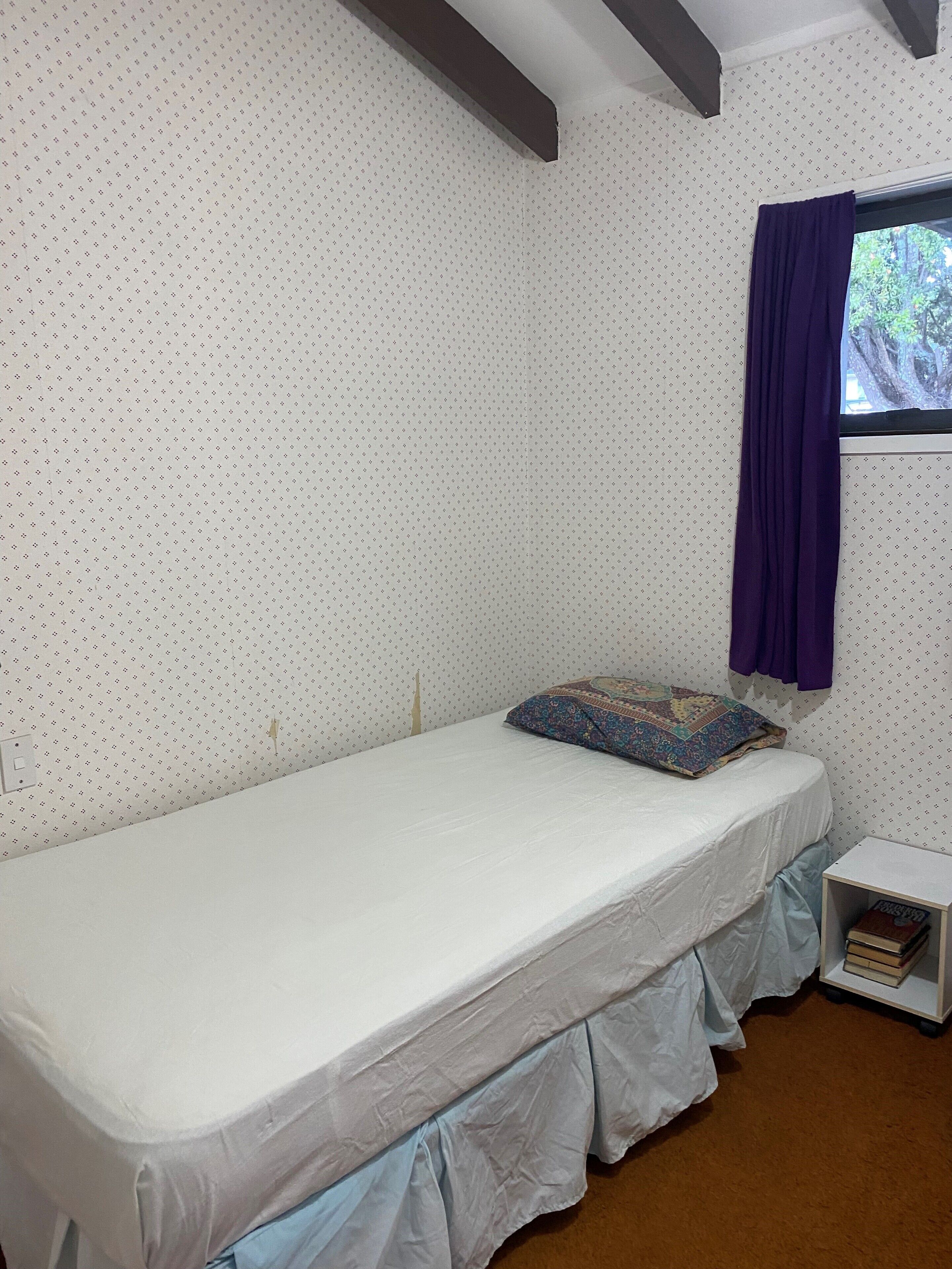 A single bed with a white mattress and a blue pillow, a window with purple curtains, and a bookshelf with books.
