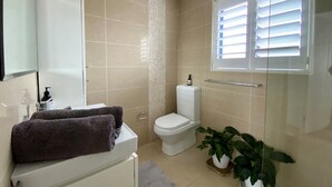 Ein Badezimmer mit beigen Fliesen, einer weißen Toilette, einer Duschkabine aus Glas, einem Handtuch auf einem weißen Schrank und zwei Zimmerpflanzen.