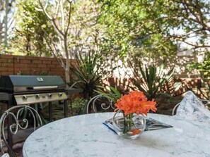 A round marble table with a clear glass vase containing orange flowers, a grill in the background, and a brick wall.