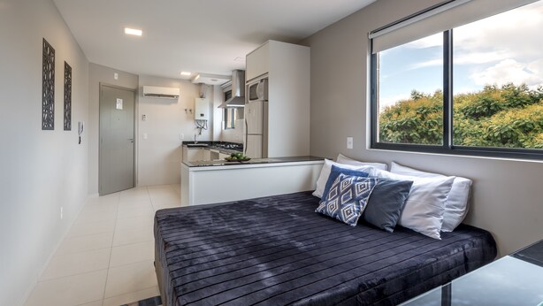A modern bedroom with a bed, a window with a view of greenery, and a kitchenette in the background. - Pleione Residence (Curitiba)