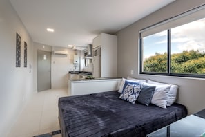 A modern bedroom with a bed, a window with a view of greenery, and a kitchenette in the background. - Pleione Residence (Curitiba)