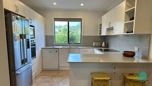 Una cocina moderna con armarios blancos, horno empotrado, refrigerador de acero inoxidable, ventana con vista a follaje y un taburete de madera.