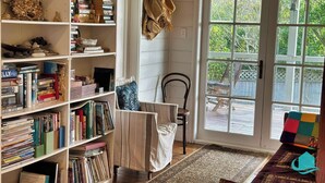 Un rincón acogedor para leer, con una silla y una estantería llena de libros, una ventana con vistas al exterior y una terraza de madera visible a través de la puerta de cristal.