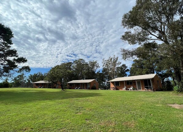 A grassy area with two brick buildings, one with a porch and a window, and another with a door and a window.