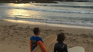 Two individuals with surfboards on a sandy beach with waves in the background.