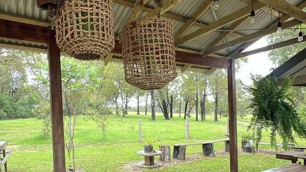 An outdoor wooden structure with a covered roof, hanging woven baskets, and a picnic table with a glass and a container on it.