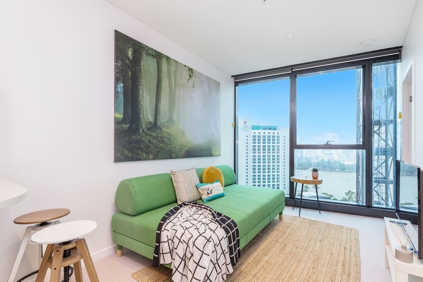 A modern living room with a green sofa, a large forest-themed painting, a view of a cityscape through large windows, and a small table with a plant.